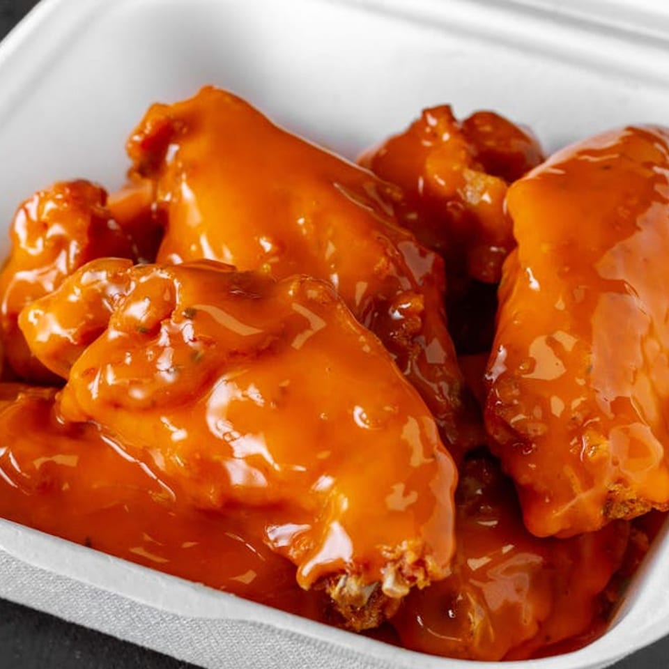 Close-up of several saucy buffalo chicken wings in a white takeout container from Liberty Pizza & Sub.