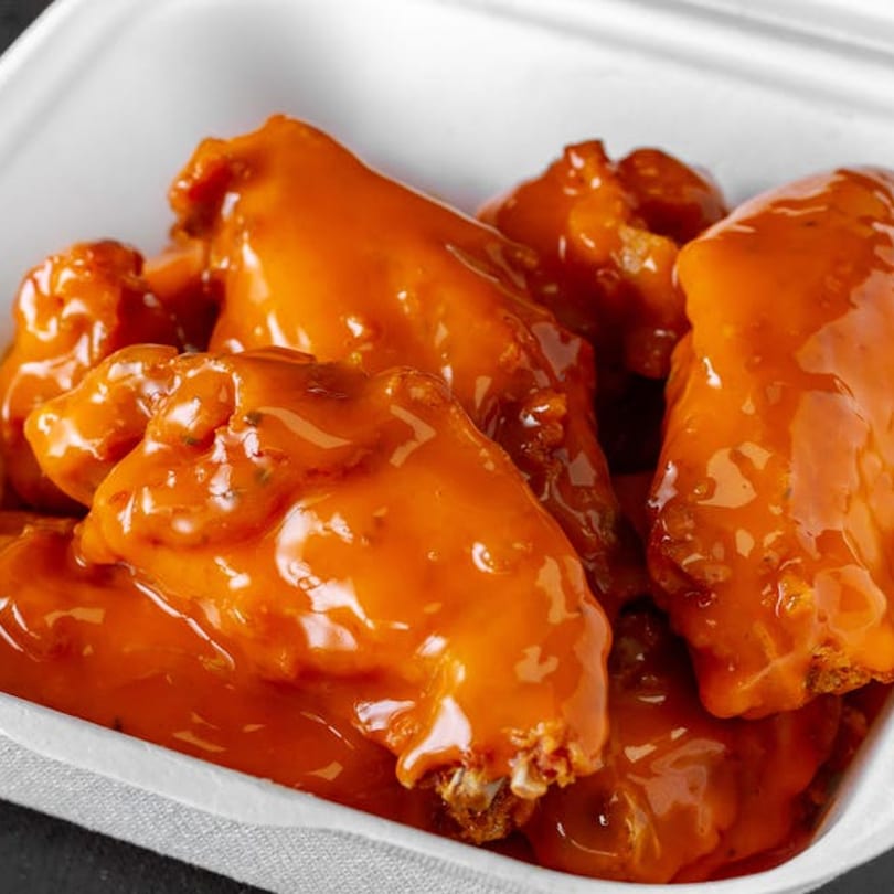Close-up of several saucy buffalo chicken wings in a white takeout container from Liberty Pizza & Sub.
