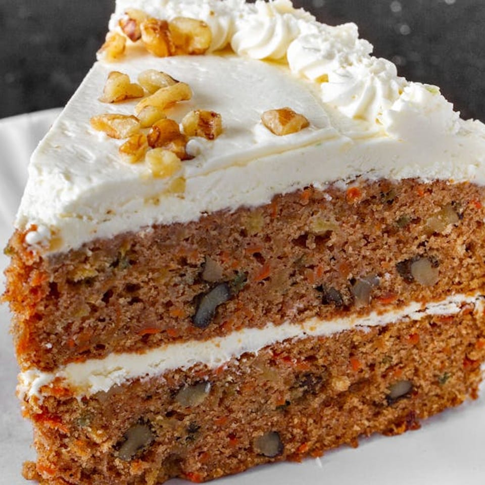 A slice of carrot cake with white frosting, topped with walnuts, on a white plate.